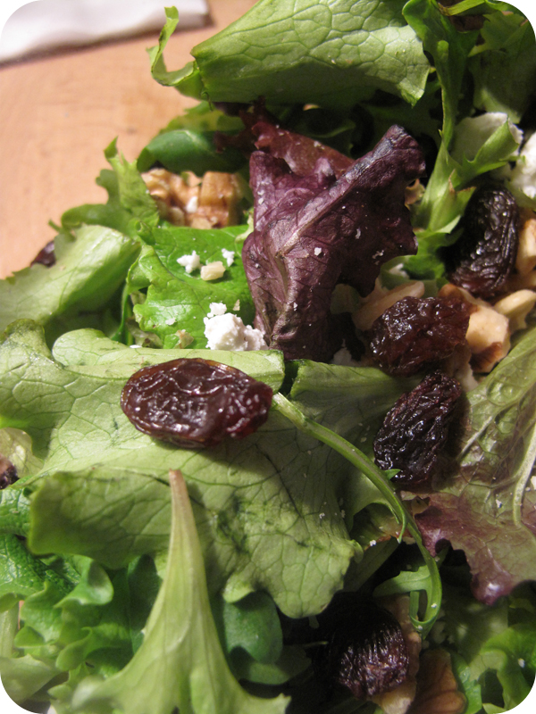 Close-up of a mixed green salad with purple lettuce, dried dark fruit, crumbled cheese, and nuts